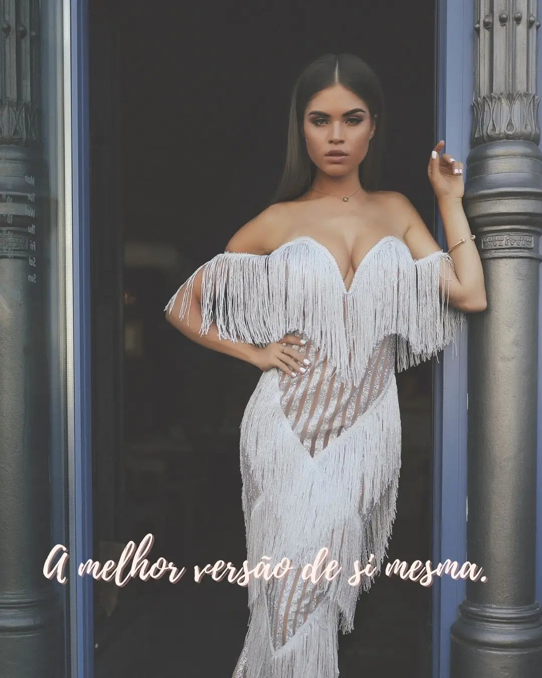 Linda mulher com vestido branco artesanal com babados, encostada em uma coluna de mármore, olhando fixamente para a camera.